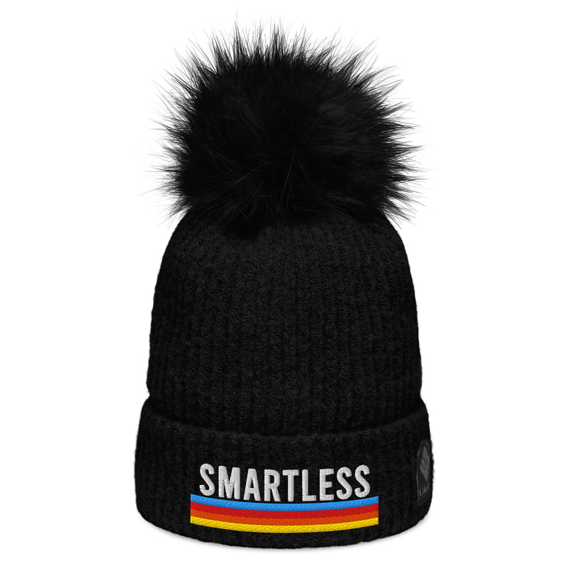 A black knit pom-pom beanie featuring the word 'SMARTLESS' embroidered in bold white capital letters on the front, above a multicolored stripe pattern in shades of blue, red, and yellow. The beanie has a ribbed texture, a folded cuff, and a black fluffy pom-pom on top.