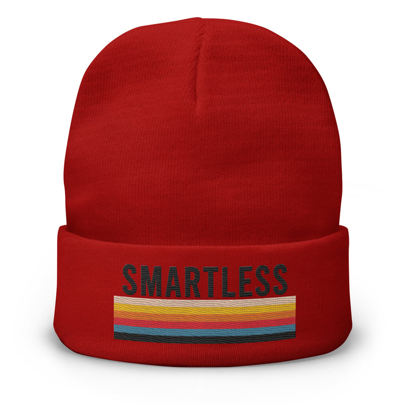 A red knit beanie with the word 'SMARTLESS' embroidered in black capital letters on the front, above a horizontal multicolored stripe pattern in shades of yellow, orange, red, and blue. The beanie is displayed against a plain white background.