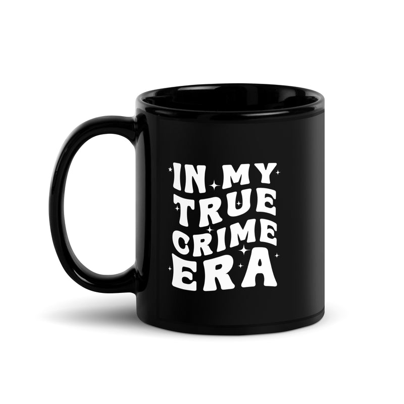 A black glossy coffee mug with a white design on the front that reads 'IN MY TRUE CRIME ERA' in a playful, retro-style font, accented with small star-like sparkles. The handle is on the left side.