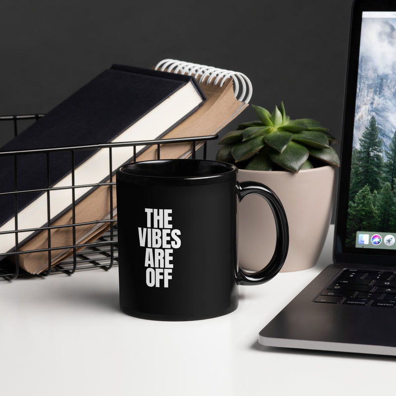 A black glossy 11 ounce coffee mug featuring bold white text that reads 'THE VIBES ARE OFF.' The mug is placed on a white desk next to a wire basket with books, a potted succulent, and a laptop, creating a stylish office setting.