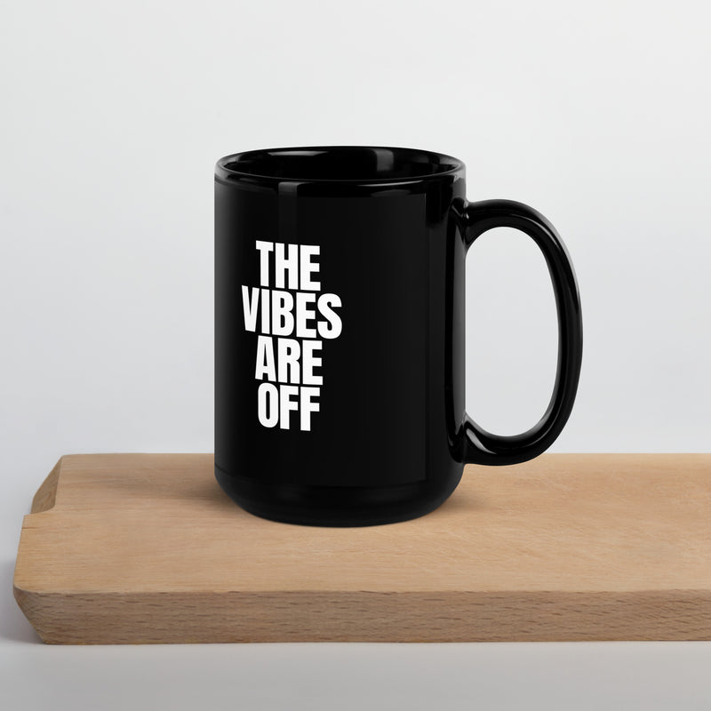 A black glossy 15 ounce coffee mug with the phrase 'THE VIBES ARE OFF' in white capital letters, positioned on a wooden surface. The mug is extra large and has a curved handle on the right side, standing out against a clean, minimalist background.