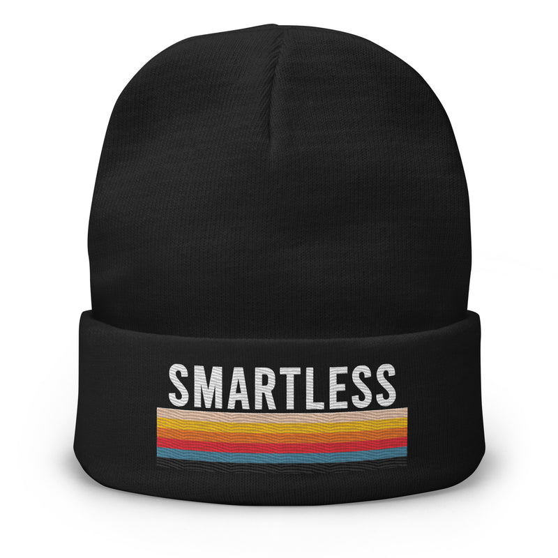 A black knit beanie with the word 'SMARTLESS' embroidered in bold white capital letters on the front, above a horizontal multicolored stripe pattern in shades of yellow, orange, red, and blue. The beanie is displayed against a plain white background.