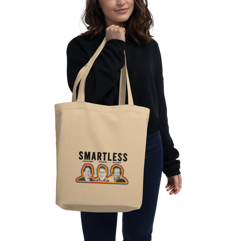 Woman holding a beige tote bag featuring the word Smartless and three illustrated faces of the hosts.