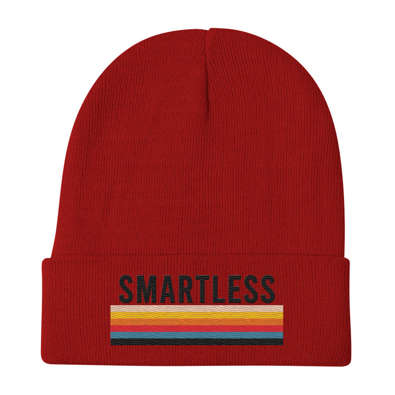 A red knit beanie with the word 'SMARTLESS' embroidered in black capital letters above a multicolored stripe pattern, displayed flat against a plain white background. The design is centered on the folded cuff of the beanie.