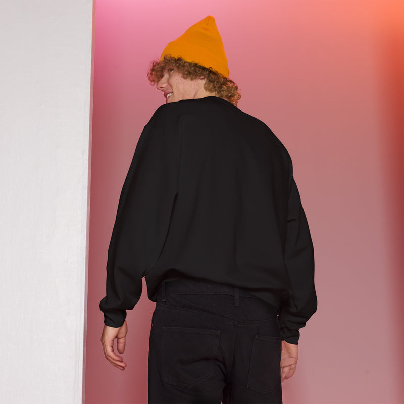 A man wearing the black crewneck sweatshirt, viewed from the back. He pairs it with black jeans and a bright yellow beanie, standing in a setting with warm pink and orange lighting.