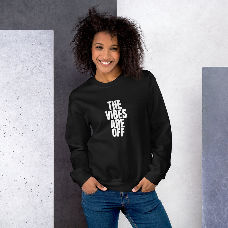 A woman wearing a black crewneck sweatshirt with bold white text that reads 'THE VIBES ARE OFF' in a stacked design. She is smiling confidently with her hands tucked into the pockets of her blue jeans. The background features a modern, geometric pattern in shades of gray and white.