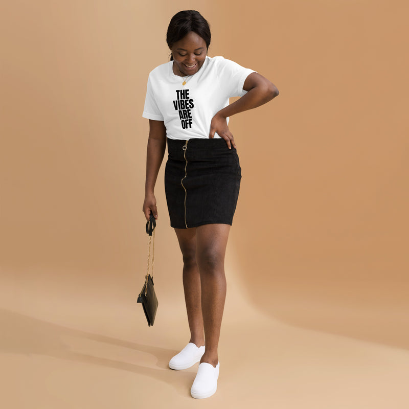 Another woman wearing the white 'THE VIBES ARE OFF' t-shirt, paired with a black skirt with gold zipper details and white slip-on sneakers, . She is smiling while adjusting her outfit, standing against a warm beige background.