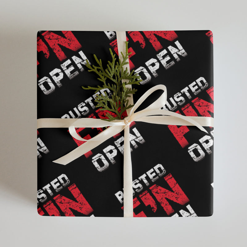 A wrapped gift with black paper with 'BUSTED F'N OPEN' repeated in red and white, tied with a cream ribbon and adorned with a small green sprig.